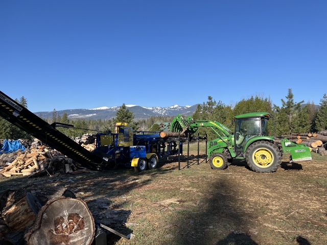 Firewood processor working alongside a tractor with a grapple loader, snowy peaks behind