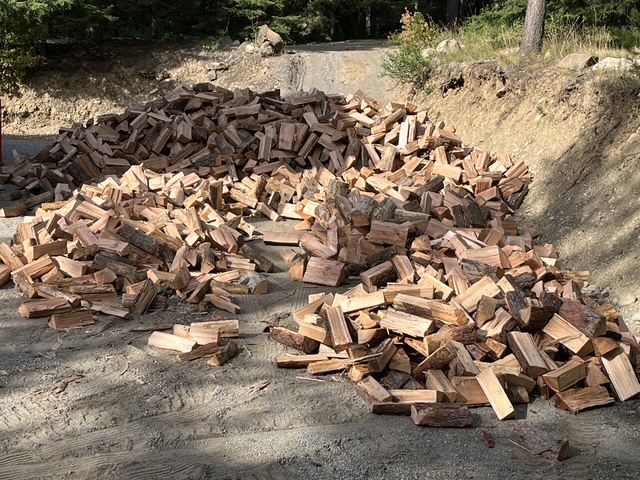 Two piles of split firewood on a gravel driveway in the trees