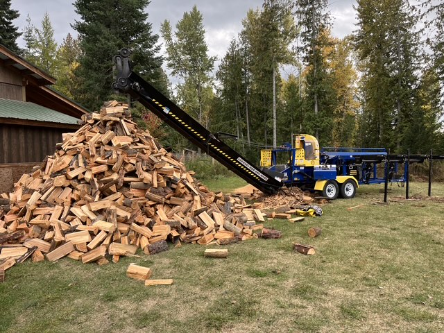 Processor conveying split firewood into a tall pile beside a cabin in the fall